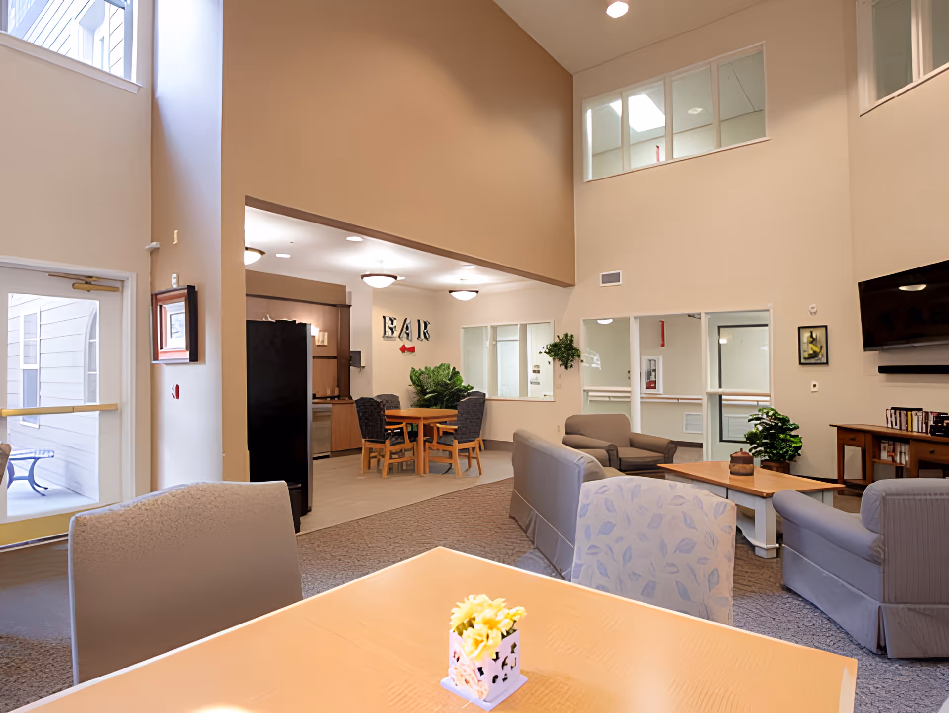 A spacious senior living common area with high ceilings and large windows. The room features comfortable seating including sofas and armchairs arranged around a coffee table. There is a dining area with a table and chairs near a small bar area. The walls are painted in neutral tones, and there are plants and framed pictures decorating the space. A television is mounted on the wall above a wooden cabinet with books and media.