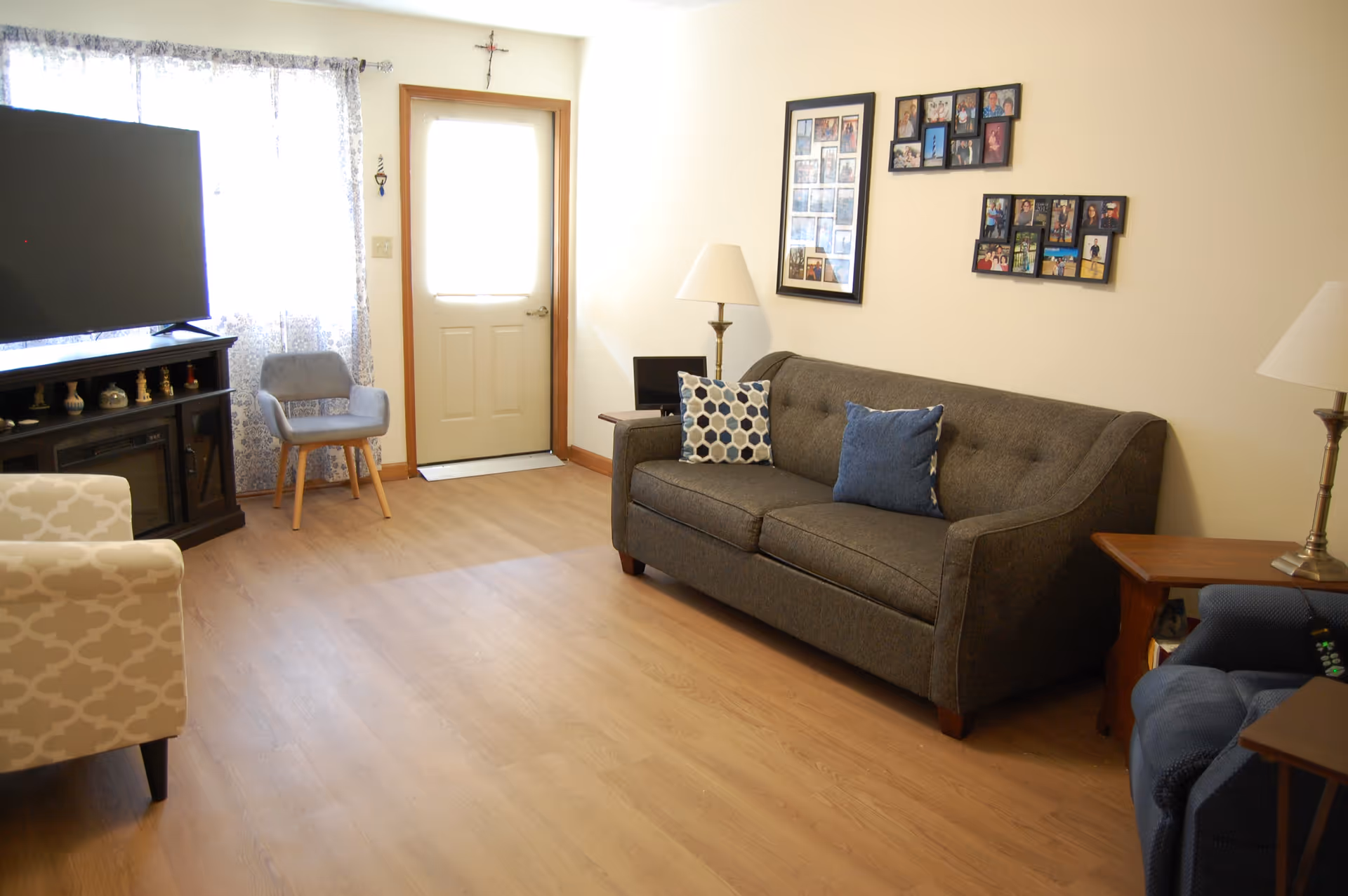 A cozy living room with a gray couch adorned with two decorative pillows, a blue armchair, a beige patterned armchair, a wooden side table with a lamp, a TV on a dark wooden stand, and a door with a window letting in natural light. Family photo collages hang on the wall above the couch.