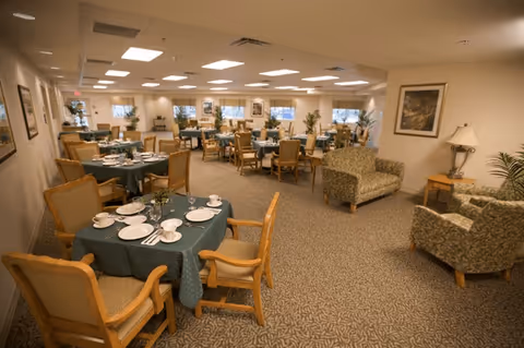 A spacious dining room with multiple round tables covered with green tablecloths, each set with white plates, cups, and silverware. Wooden chairs with cushioned seats surround the tables. The room is well-lit with ceiling lights and has several windows along the far wall. There are also a couple of upholstered armchairs and a small side table with a lamp in one corner. The walls are decorated with framed artwork and plants are placed around the room.