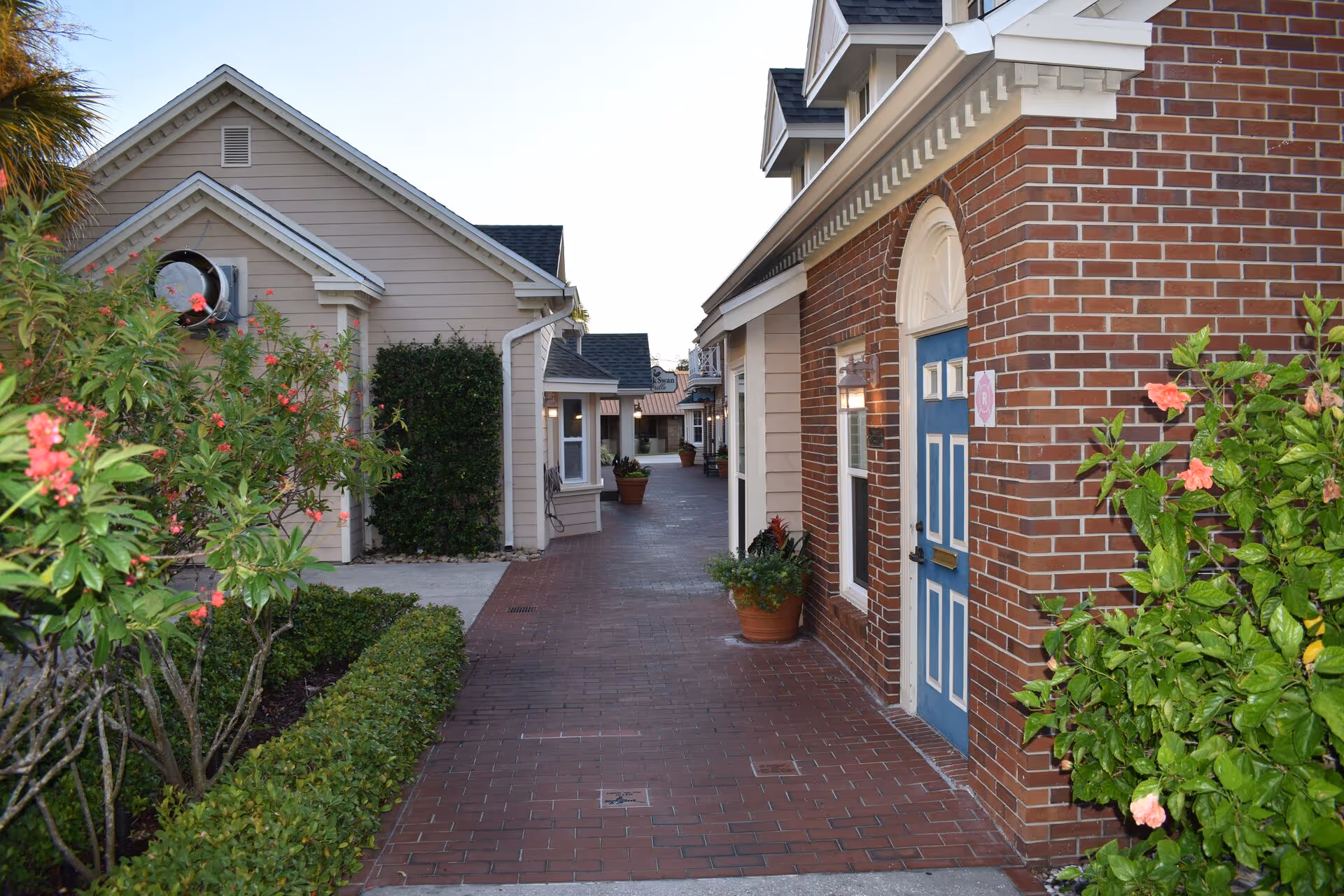 A brick-paved walkway between buildings at a senior living facility, with a brick building featuring a blue door on the right and beige siding buildings on the left. There are potted plants and flowering bushes along the walkway.