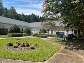 Outdoor courtyard area at Lancaster Grove Senior Living featuring a circular flower bed with various plants, surrounded by a well-maintained lawn and paved walkways. There are benches and chairs under a tree with pink blossoms, and the building with white siding and multiple windows encloses the courtyard. Trees and greenery are visible in the background under a blue sky with some clouds.