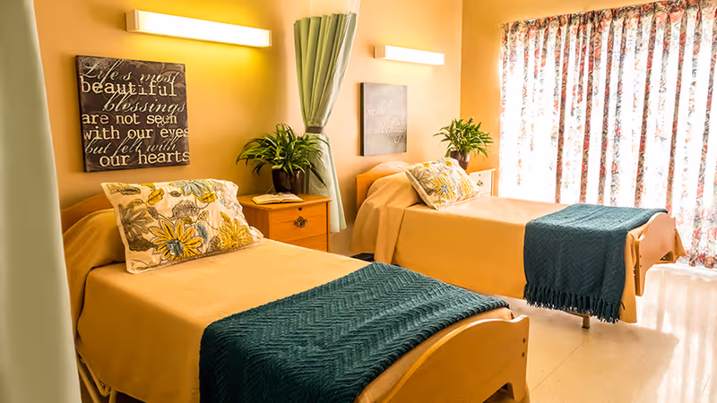 A bright and cozy nursing center bedroom with two single beds, each covered with a beige blanket and a dark green throw. Each bed has a floral pillow and a wooden headboard. Between the beds is a wooden nightstand with a potted plant and a book. The walls are painted warm yellow and decorated with framed inspirational quotes. A window with floral curtains allows natural light to fill the room.