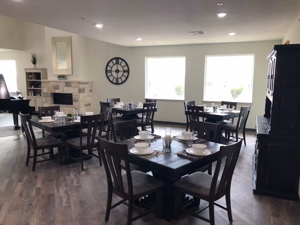 A dining room with several dark wooden tables and chairs set with white bowls, plates, glasses, and silverware. The room has large windows letting in natural light, a stone fireplace with a mirror above it, a large wall clock, and a dark wooden cabinet on the right side. The floor is wood, and there is a piano visible in the background.
