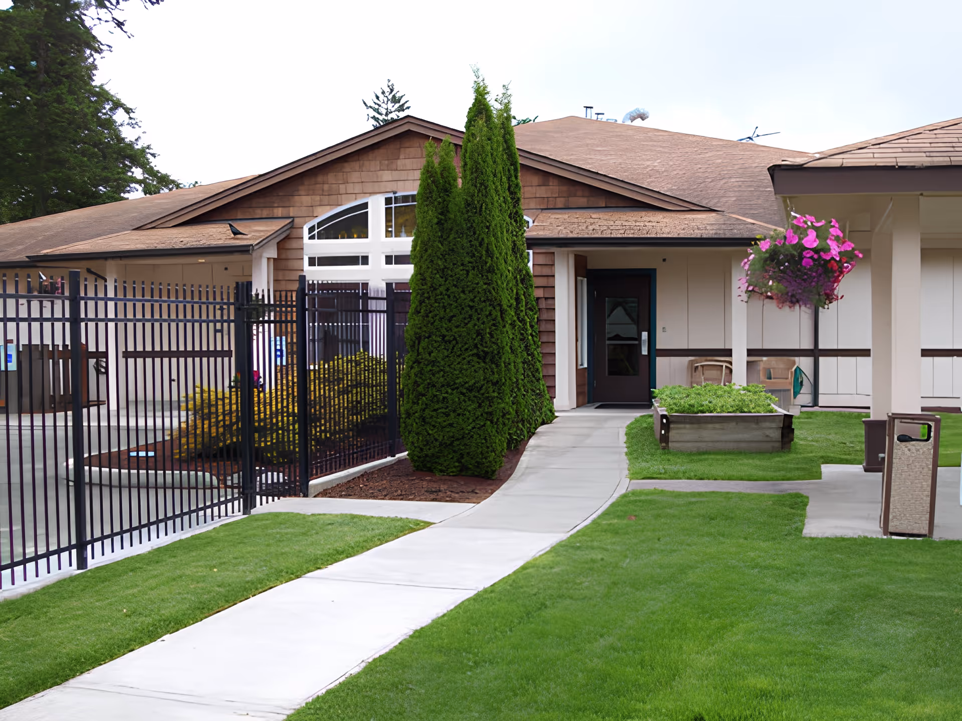 Exterior view of a single-story building with brown shingle siding and a sloped roof. A concrete pathway leads to a dark-colored door entrance. There are tall green shrubs along the pathway, a black metal fence on the left, a hanging basket with pink flowers on the right, and a small garden bed with greenery near the entrance. The lawn is well-maintained and green.