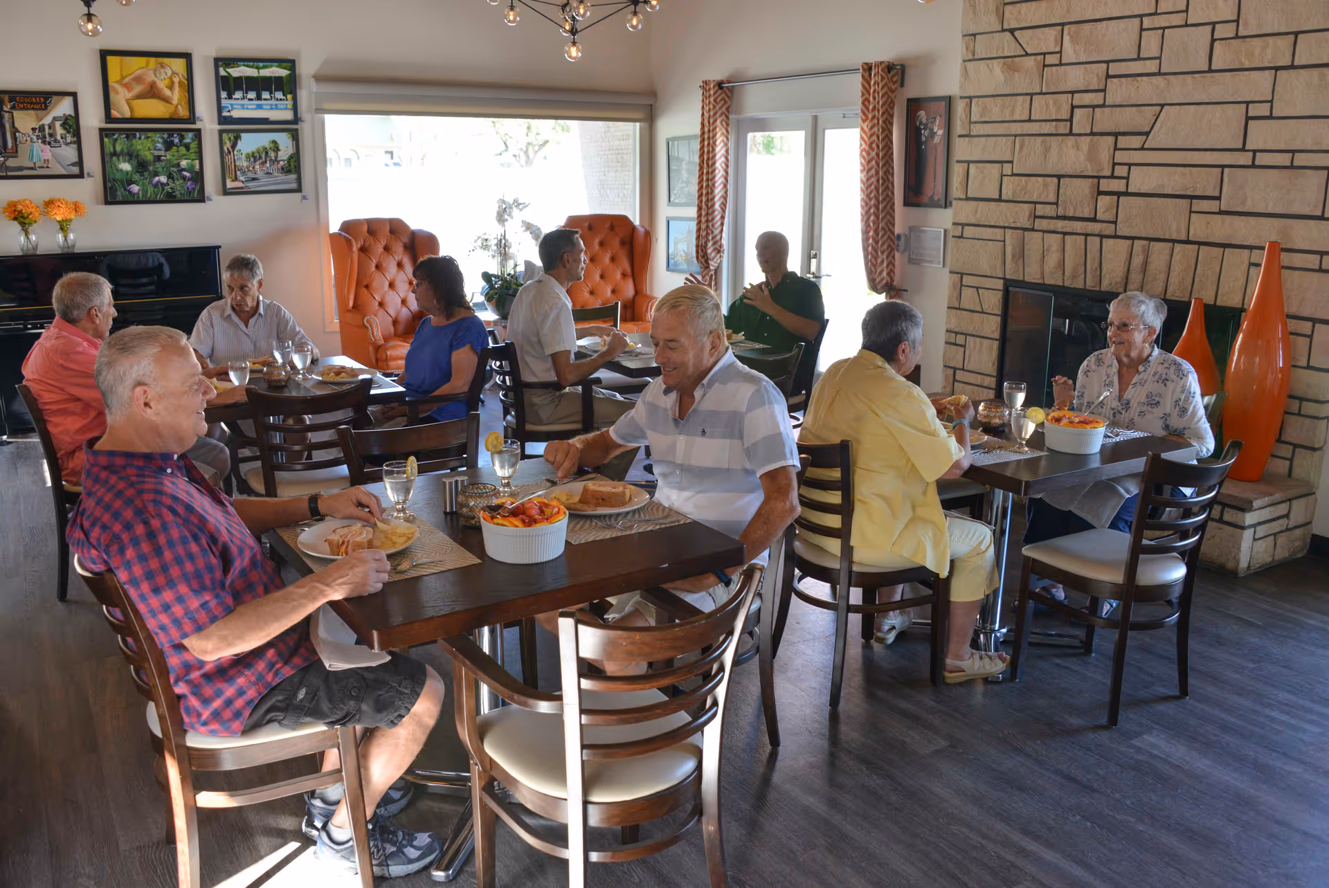 A group of elderly people sitting at tables in a well-lit dining room, enjoying a meal together. The room features wooden chairs and tables, a stone fireplace with orange decorative vases, framed pictures on the walls, and large windows letting in natural light.