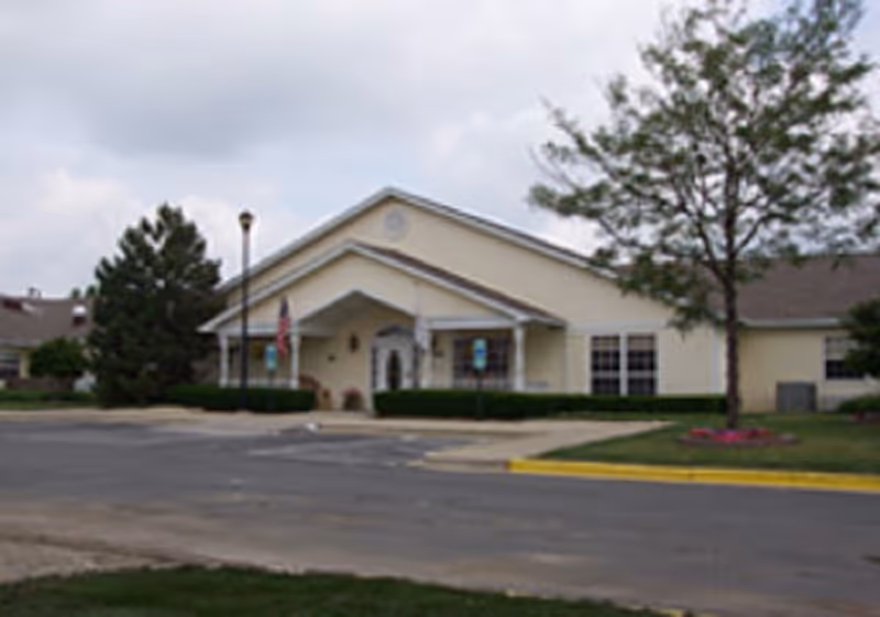 Exterior view of a single-story building with a peaked roof, beige walls, and several windows. There is a paved driveway and parking area in front, a tree with some flowers planted around its base, and a flagpole near the entrance.
