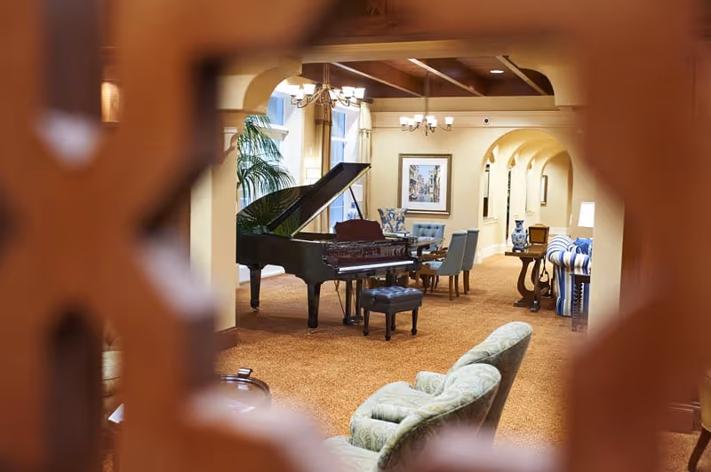 View through a decorative wooden screen into a spacious, warmly lit common area featuring a grand piano, several upholstered chairs, a round table with chairs, framed artwork on the walls, and arched doorways in the background.