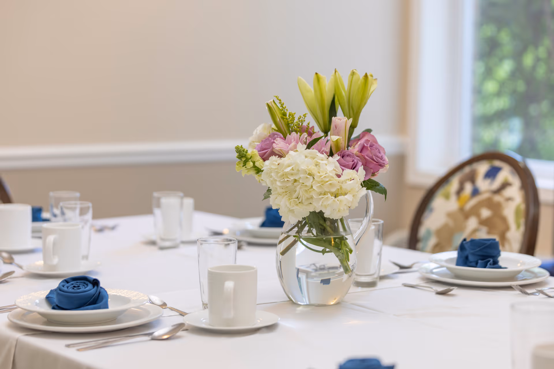 A dining table set with white plates, cups, and silverware, featuring blue cloth napkins folded in a rose shape. A clear glass pitcher with a bouquet of white, pink, and yellow flowers serves as the centerpiece. The background shows a window and a chair with a patterned cushion.