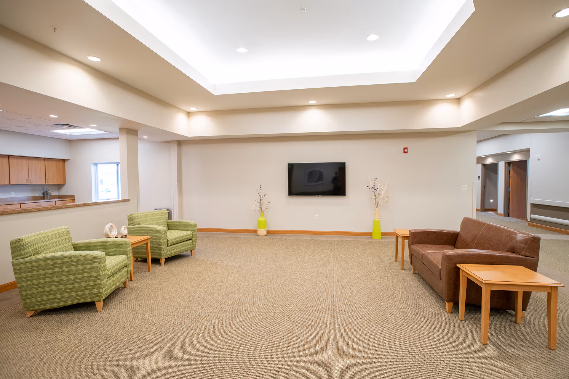 A spacious, well-lit common area with beige walls and carpeted floor. The room features two green armchairs and a small wooden side table on the left, a brown leather loveseat with two wooden side tables on the right, and a flat-screen TV mounted on the far wall. Two decorative vases with branches are placed on the floor beneath the TV. The ceiling has recessed lighting and a large central light fixture.