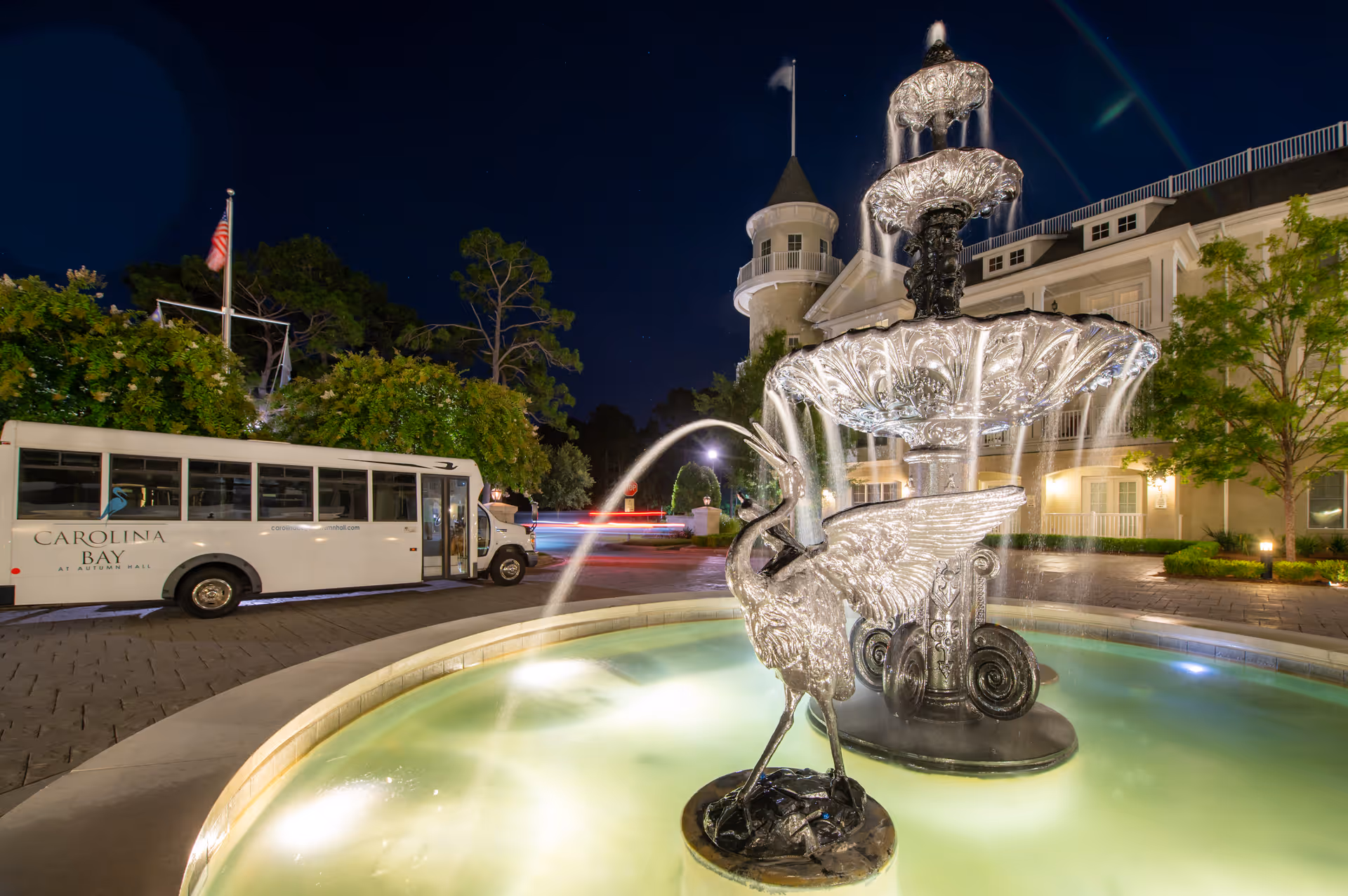 Night view of an ornate multi-tiered fountain with water flowing, featuring a bird sculpture in the foreground. Behind the fountain is a white shuttle bus with the text 'Carolina Bay at Autumn Hall' parked on a paved driveway. The background shows a large building with a turret and lit windows, surrounded by trees and landscaping.
