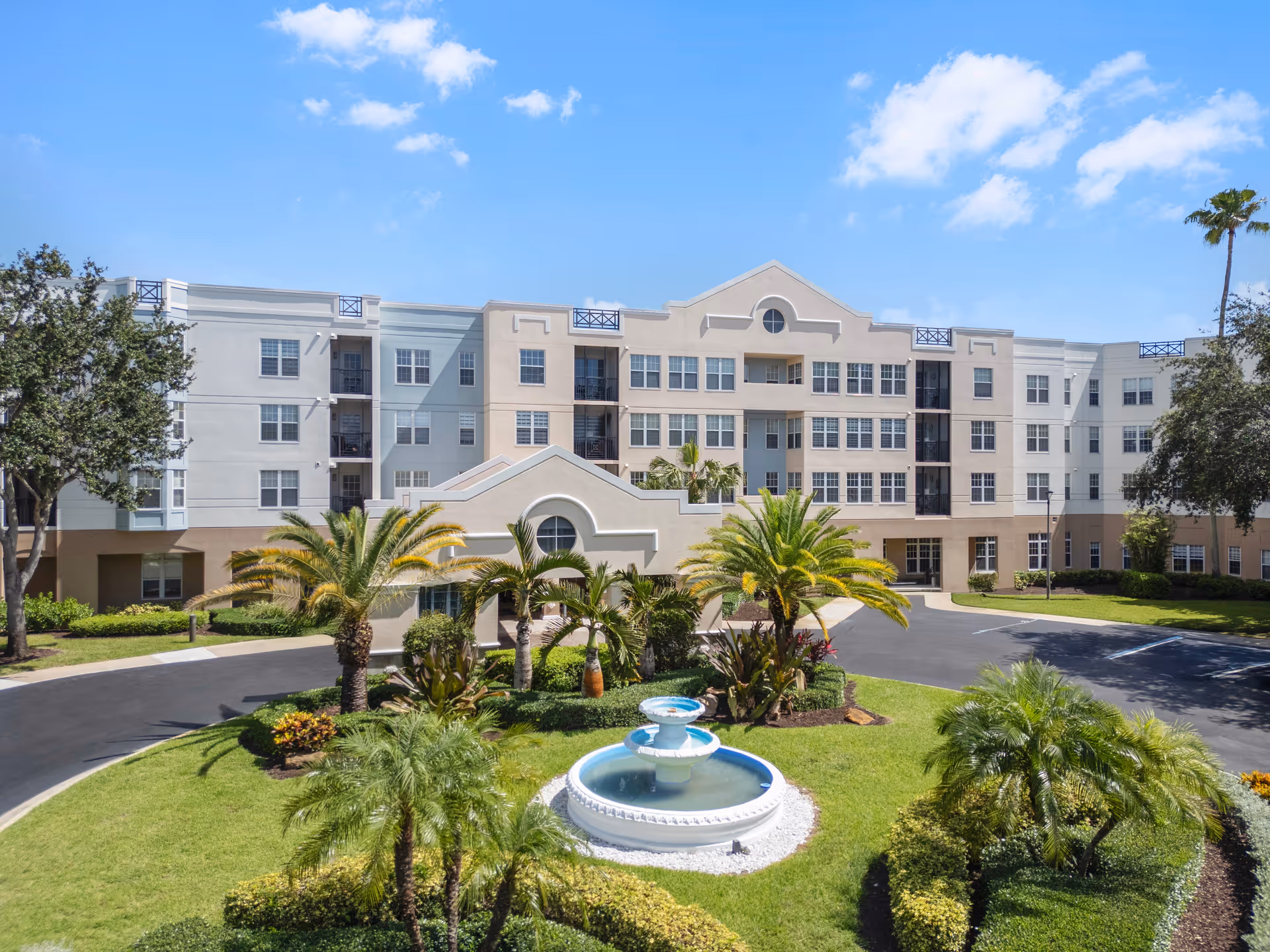 Exterior view of a multi-story senior living facility building with a circular driveway and landscaped garden featuring palm trees and a decorative water fountain in the center under a blue sky with some clouds.