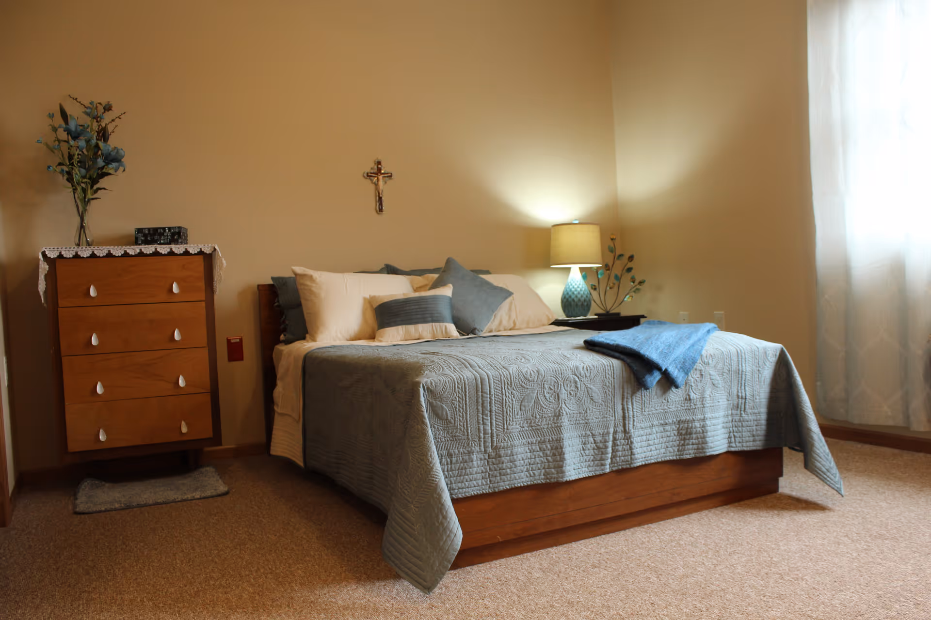 A cozy bedroom with a wooden bed covered in a blue quilt and multiple pillows. A blue throw blanket is draped over the bed. To the left of the bed is a wooden chest of drawers with a lace doily and a vase with flowers on top. A small rug is placed in front of the chest. A lamp with a blue base and a beige shade sits on a nightstand to the right of the bed. A crucifix hangs on the wall above the bed. Light filters softly through a sheer curtain on the right side of the room.