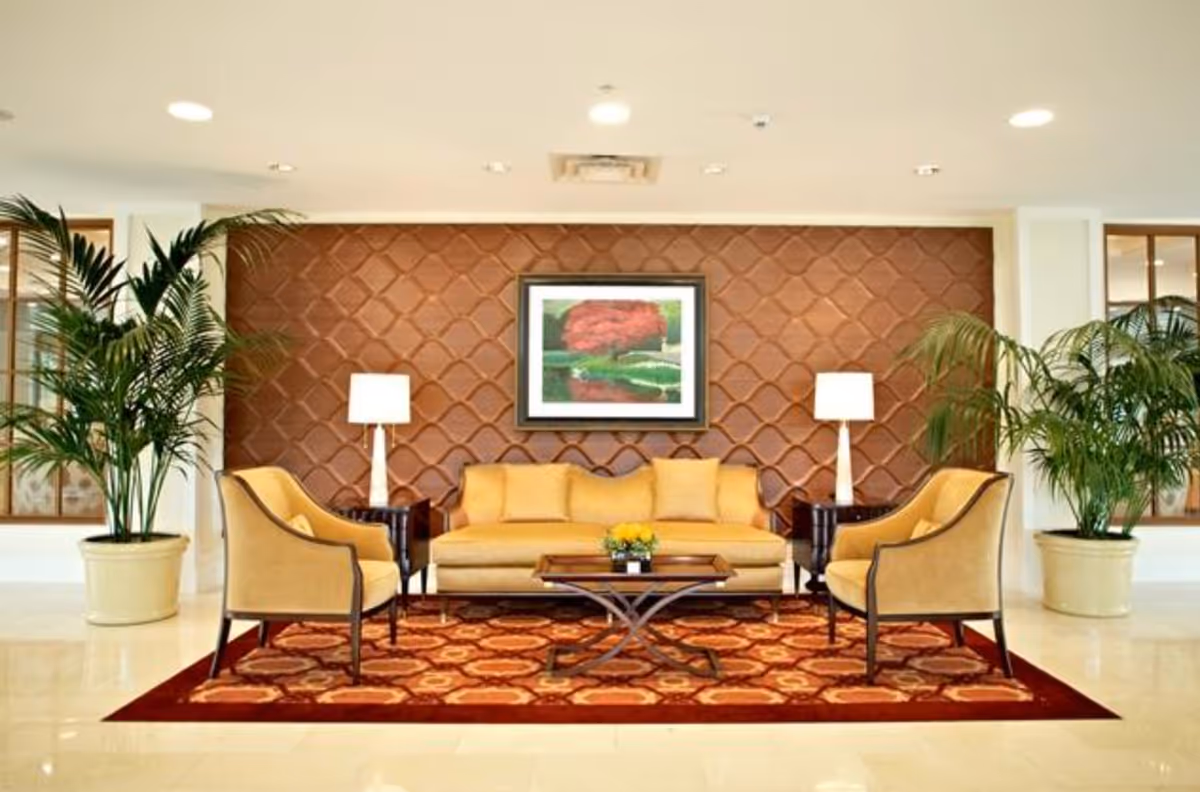 A well-lit living room area with a yellow sofa and two matching armchairs arranged around a wooden coffee table on a patterned red and orange rug. Two white table lamps sit on dark wooden side tables on either side of the sofa. The back wall features a textured brown design with a framed painting of a red tree above the sofa. Large potted plants are placed on either side of the seating area.