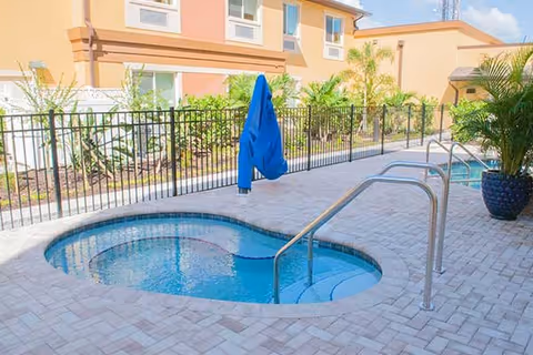 Outdoor area with a small round pool or hot tub surrounded by a tiled patio, metal handrails, a closed blue umbrella, and a building with windows and plants in the background.