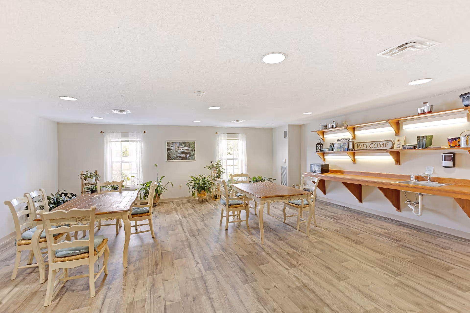 A bright and spacious common area with wooden floors, two wooden tables surrounded by chairs with cushions, several potted plants near two windows with sheer curtains, and a long wooden counter with shelves above it displaying books, a welcome sign, and various small items.