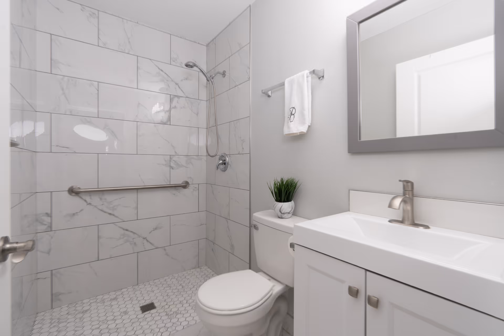 A clean, modern bathroom featuring a walk-in shower with large white marble tiles and a stainless steel grab bar. The bathroom also includes a white toilet with a small green plant on top, a white vanity with a sink and silver faucet, a rectangular mirror with a gray frame, and a towel rack holding a white towel with a decorative letter B.