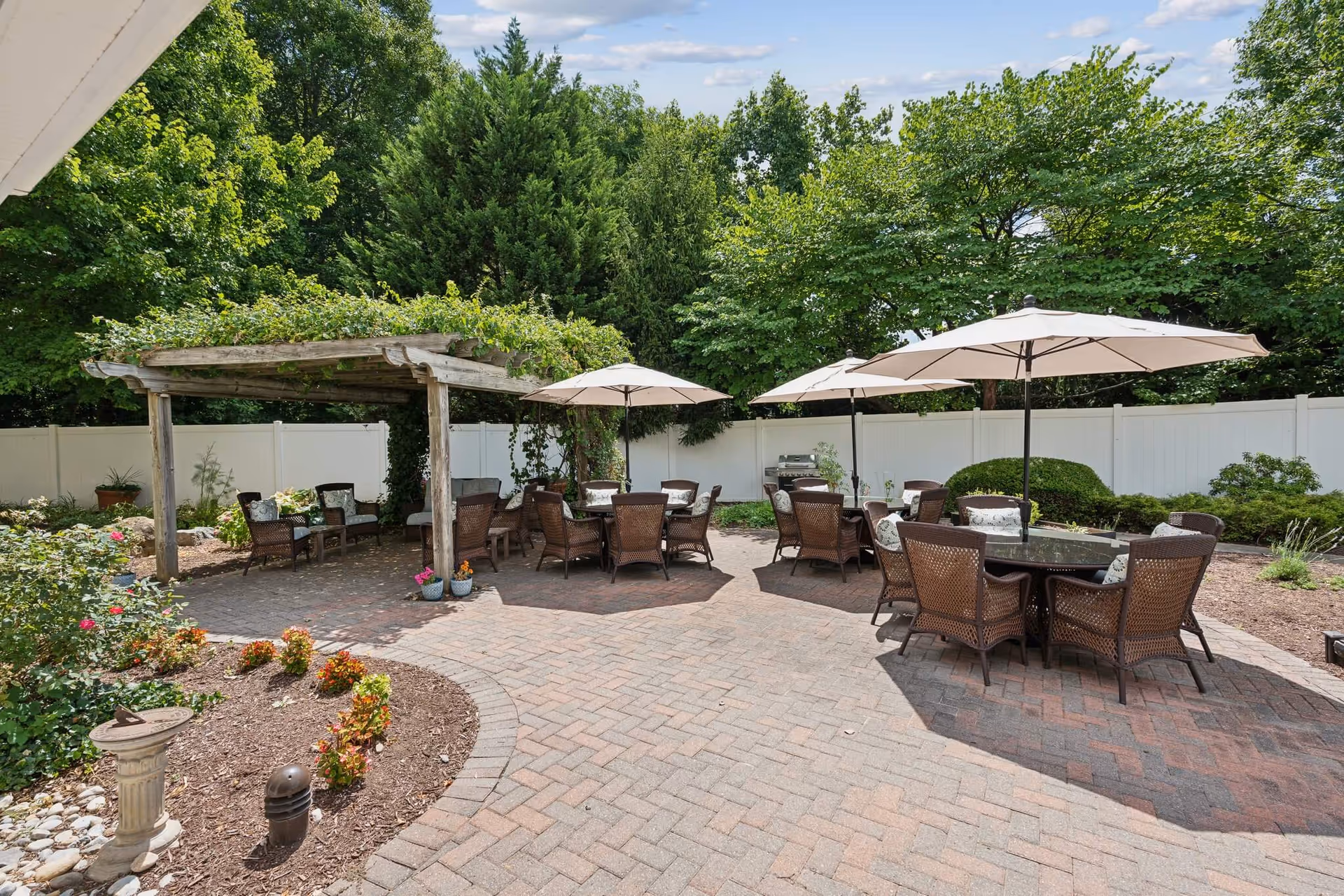 Outdoor patio area with multiple round tables surrounded by wicker chairs, each table shaded by large beige umbrellas. There is a wooden pergola covered with greenery on the left side, and various plants and flowers along the edges of the paved brick patio. A white fence and tall green trees surround the area under a partly cloudy sky.