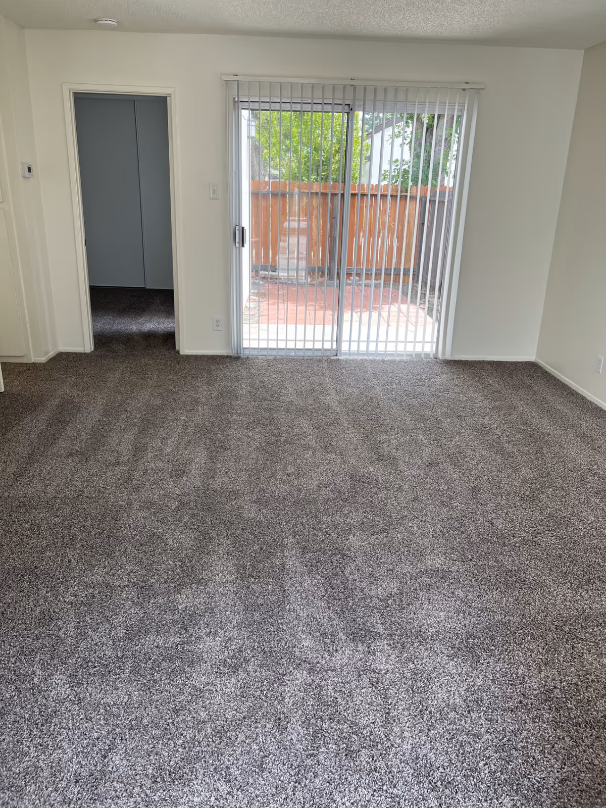 Empty room with gray carpet flooring, white walls, a sliding glass door with vertical blinds leading to an outdoor patio area with a wooden fence, and an open doorway leading to another room.