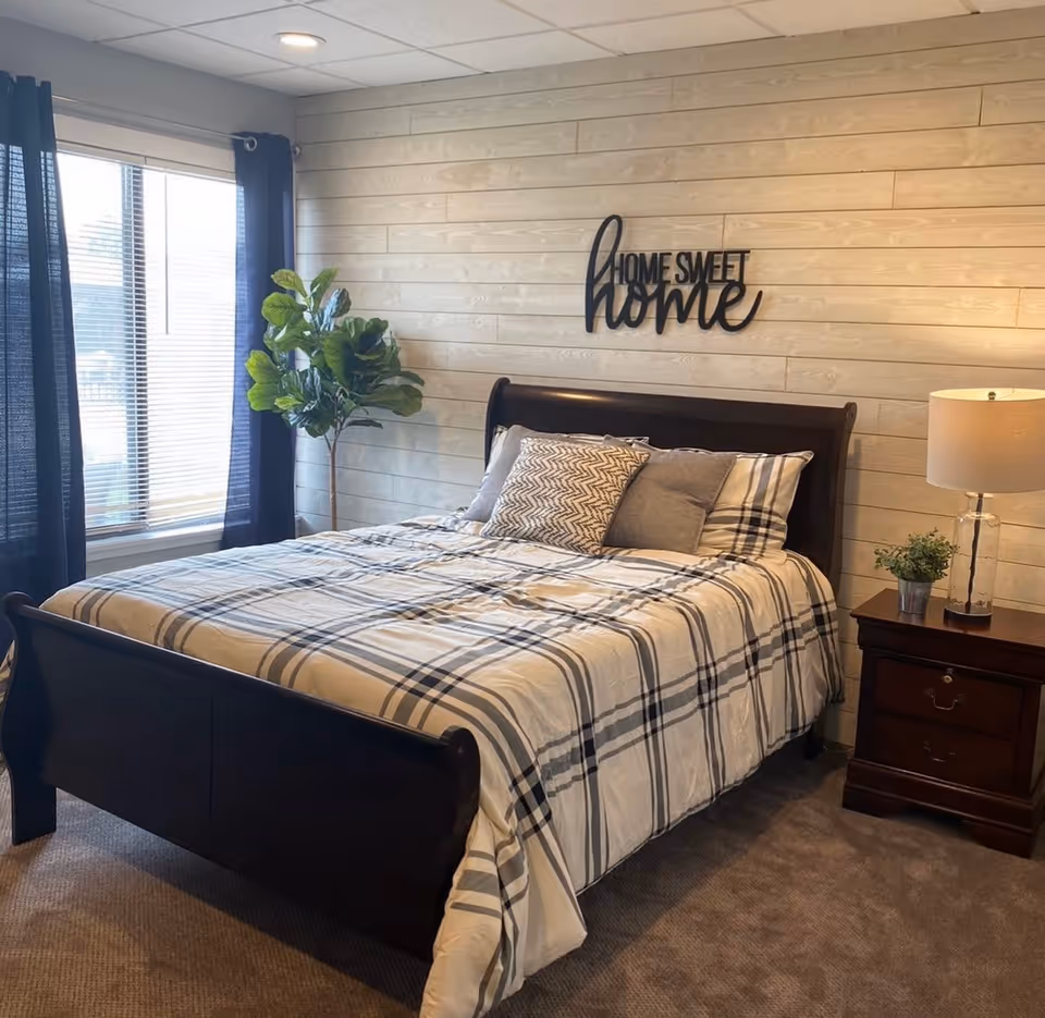 A cozy bedroom with a dark wooden bed frame and a plaid bedspread. There are three decorative pillows on the bed. A wooden nightstand with a lamp and a small potted plant is next to the bed. A large window with dark curtains allows natural light into the room. A green potted plant stands in the corner. On the wall above the bed, there is a decorative sign that reads 'Home Sweet Home'.