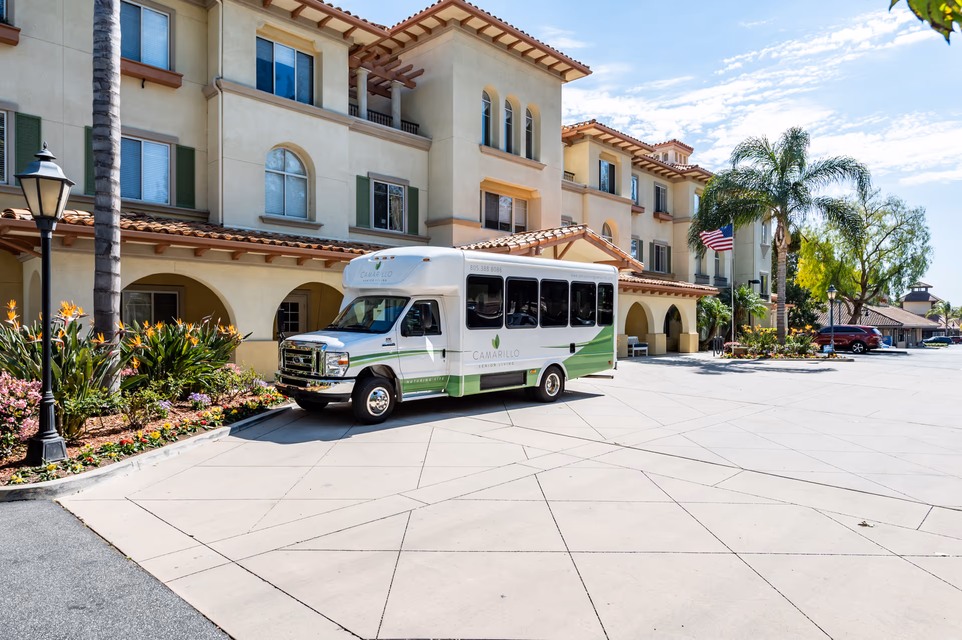 Exterior view of Camarillo Senior Living facility showing a beige multi-story building with arched windows and a tiled roof. A white and green shuttle bus with the Camarillo Senior Living logo is parked in front. There are palm trees, flowers, and a lamppost near the entrance, with a clear blue sky overhead.