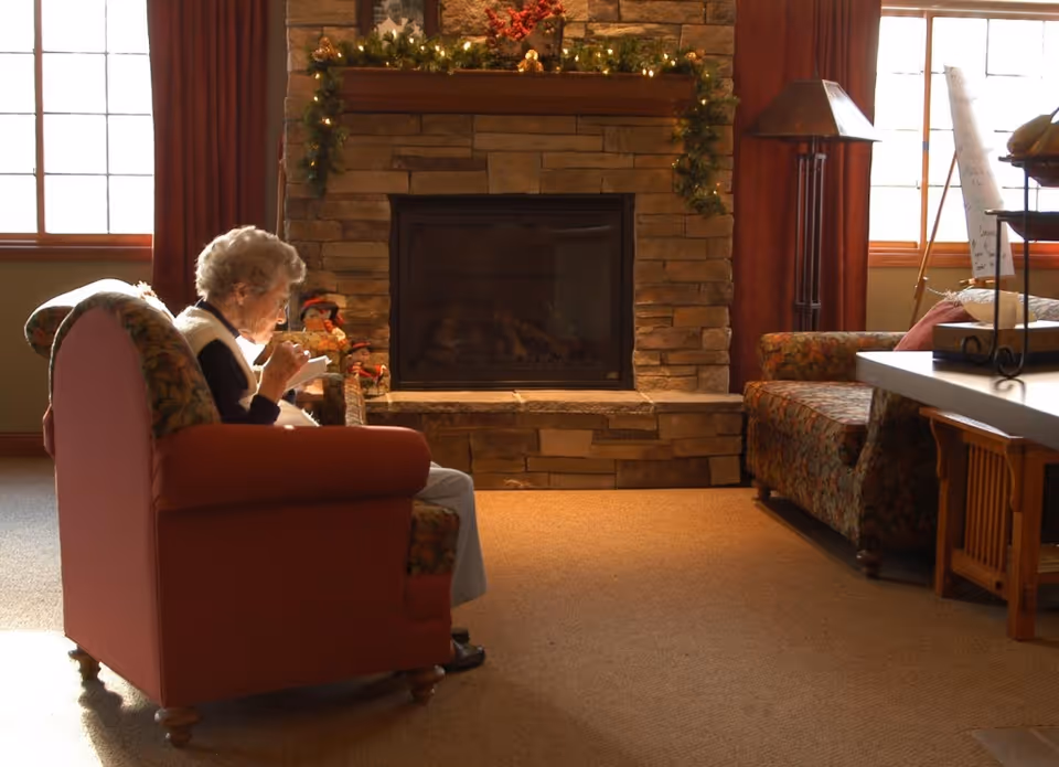 An elderly woman sitting in a cushioned armchair in a cozy living room with a stone fireplace decorated with garland and holiday ornaments. The room has large windows with red curtains, a patterned sofa, a floor lamp, and a carpeted floor.