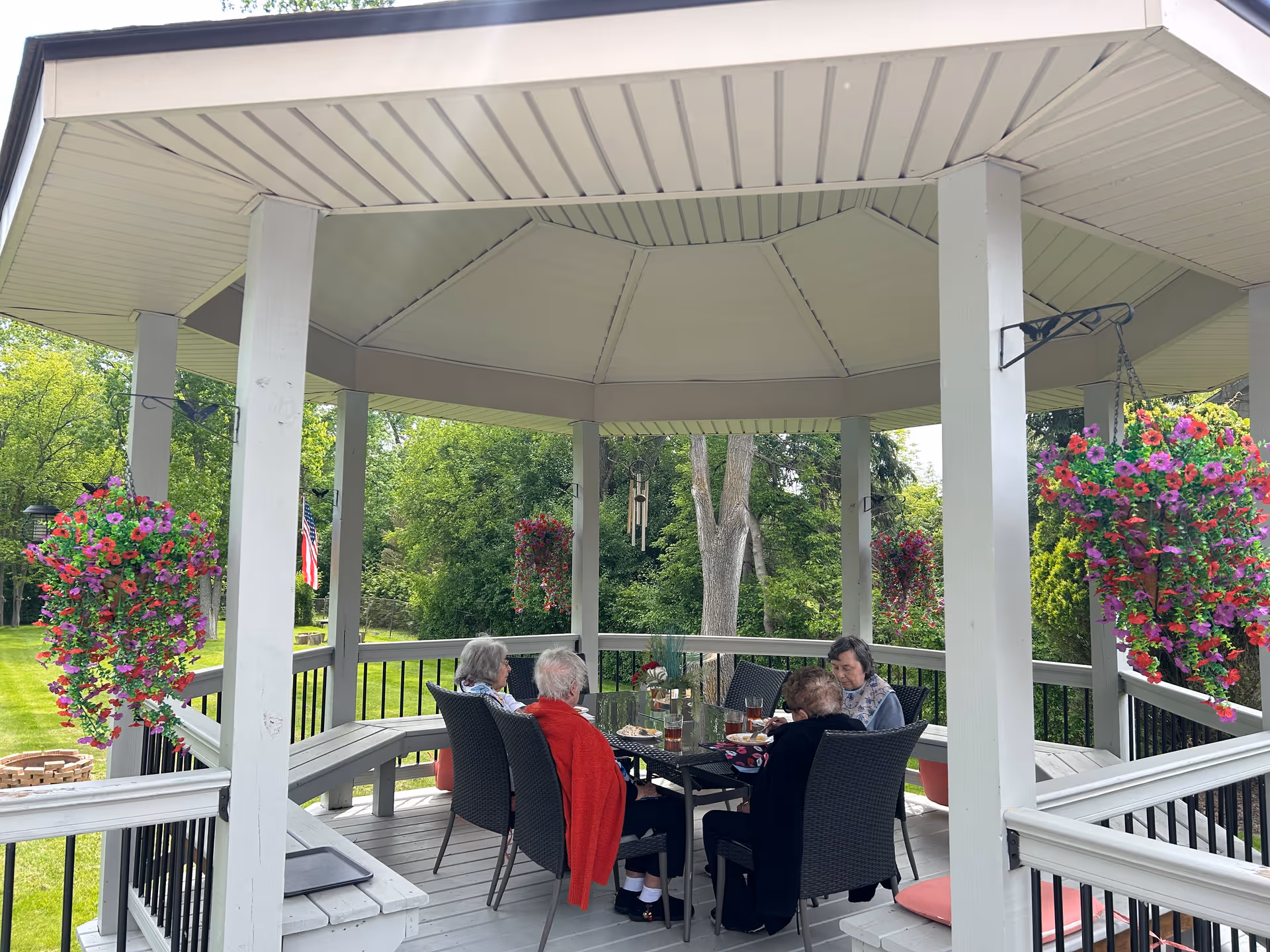 Four elderly women sitting around a glass table under a white gazebo in a garden. The gazebo has hanging baskets with vibrant pink and purple flowers. The surrounding area is lush with green trees and grass, and an American flag is visible in the background.