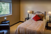 A small bedroom with a single bed covered in a beige comforter and a red pillow. There are two bedside tables, each with a lamp. A window with a view of greenery is on the left wall, and a dark armchair is positioned near the window.