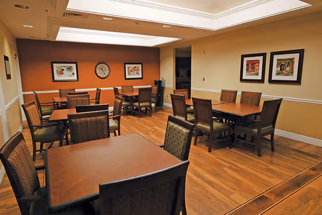 Interior view of a dining room with several wooden tables and chairs arranged neatly on a wooden floor. The walls are painted beige and orange with framed artwork and a clock hanging on them. The ceiling has recessed lighting.