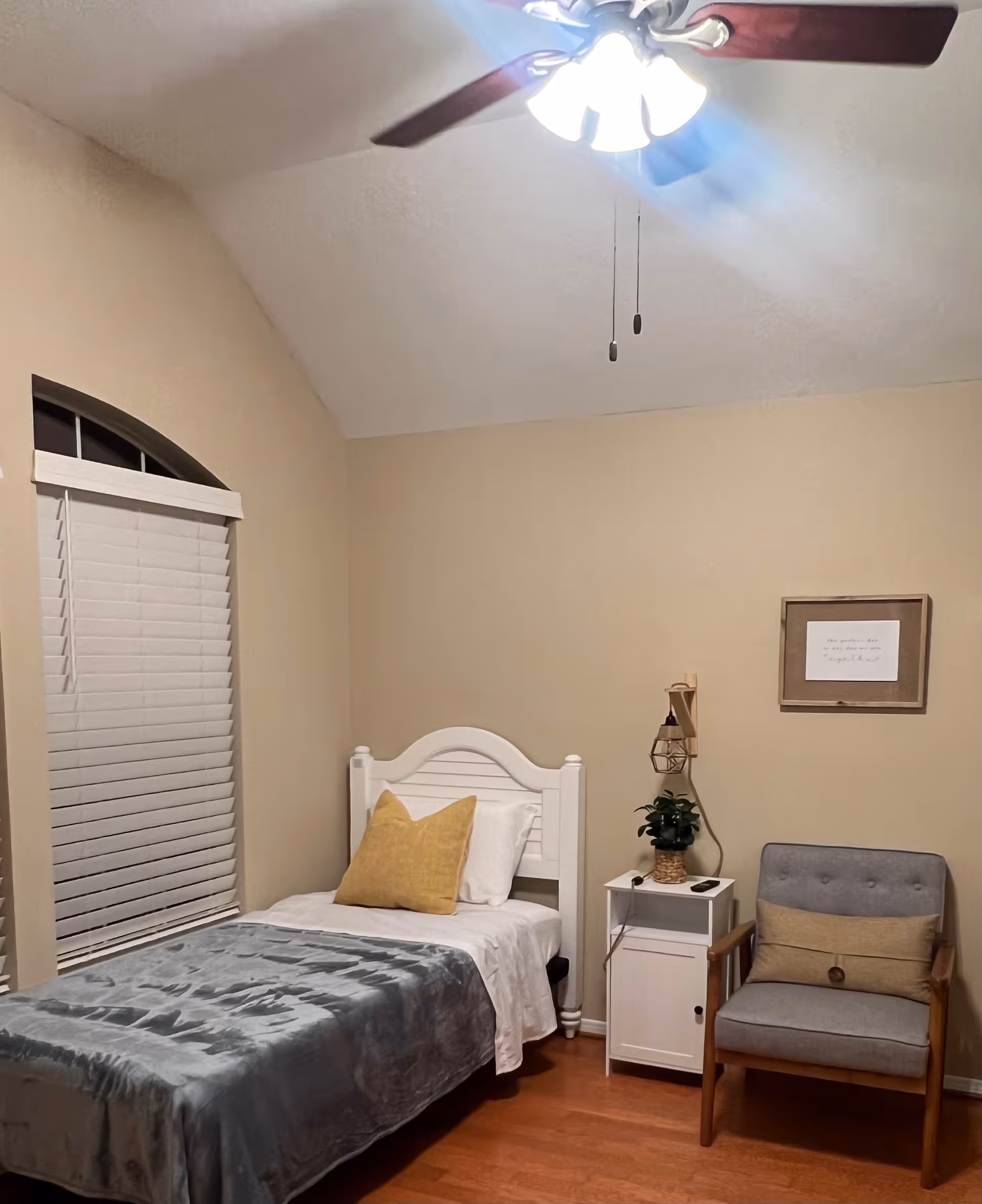 A cozy bedroom with a single white bed featuring a yellow pillow and gray blanket. Next to the bed is a white nightstand with a potted plant and a small lamp mounted on the wall above it. A gray cushioned chair with wooden arms and a beige pillow is placed beside the nightstand. The room has beige walls, a window with white blinds, and a ceiling fan with lights.