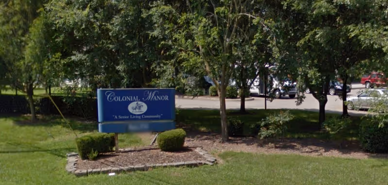 A blue Colonial Manor sign on a grassy lawn with trees