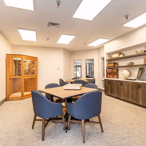 A well-lit interior room with a square table surrounded by four blue upholstered chairs. The room features a wooden cabinet with glass doors on the left, a long wooden sideboard with shelves displaying decorative items including a globe and books on the right, and a doorway leading to another area in the background. The ceiling has multiple rectangular light fixtures.
