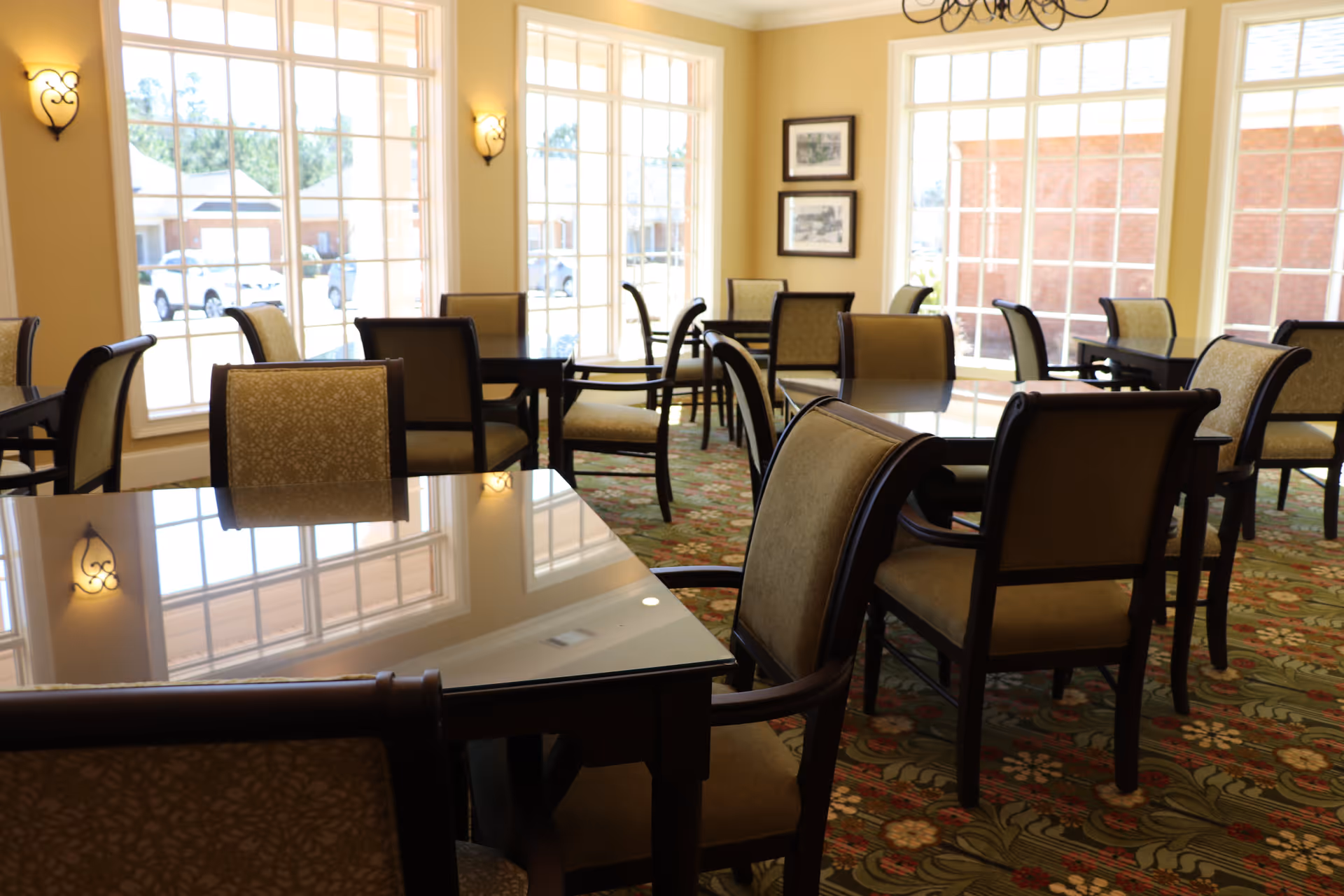 A bright dining room with multiple tables and cushioned chairs arranged neatly. Large windows allow natural light to fill the room, and wall sconces provide additional lighting. The room features a patterned carpet and framed artwork on the walls.