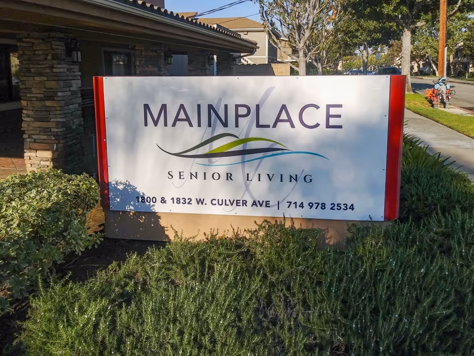 Outdoor sign for Mainplace Senior Living with the address 1800 & 1832 W. Culver Ave and phone number 714 978 2534, surrounded by greenery and a building in the background.