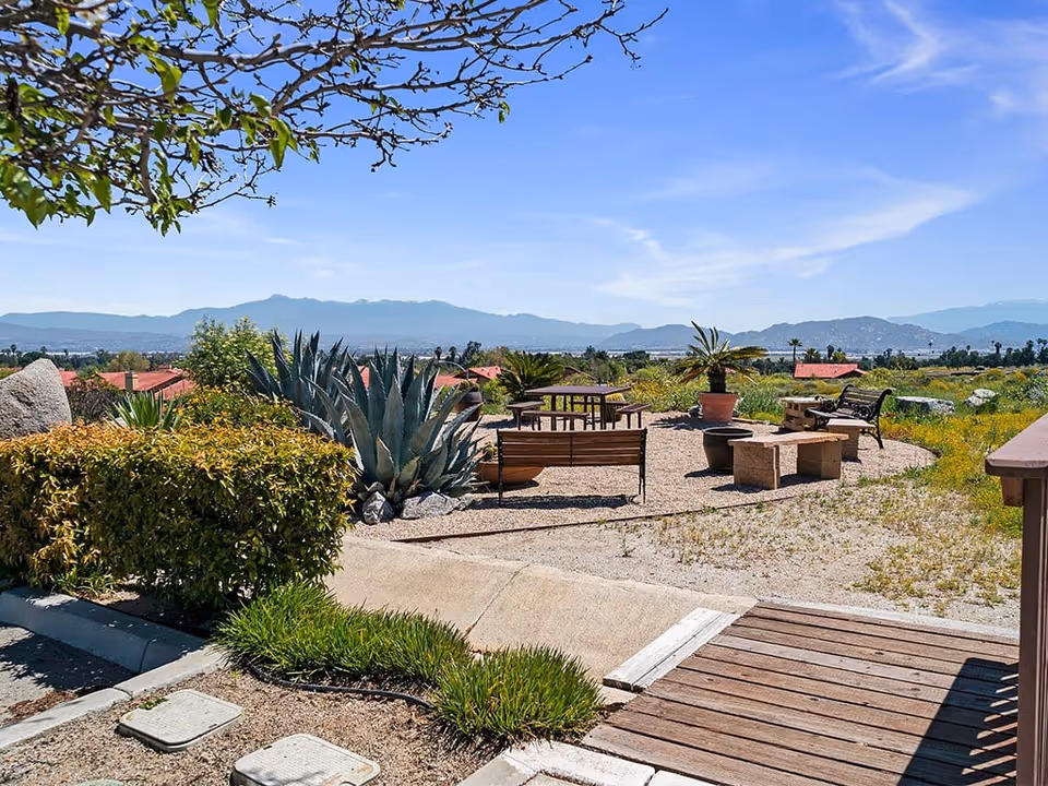 Outdoor seating area with benches, a picnic table, and potted plants surrounded by gravel and greenery, with mountains visible in the background under a clear blue sky.