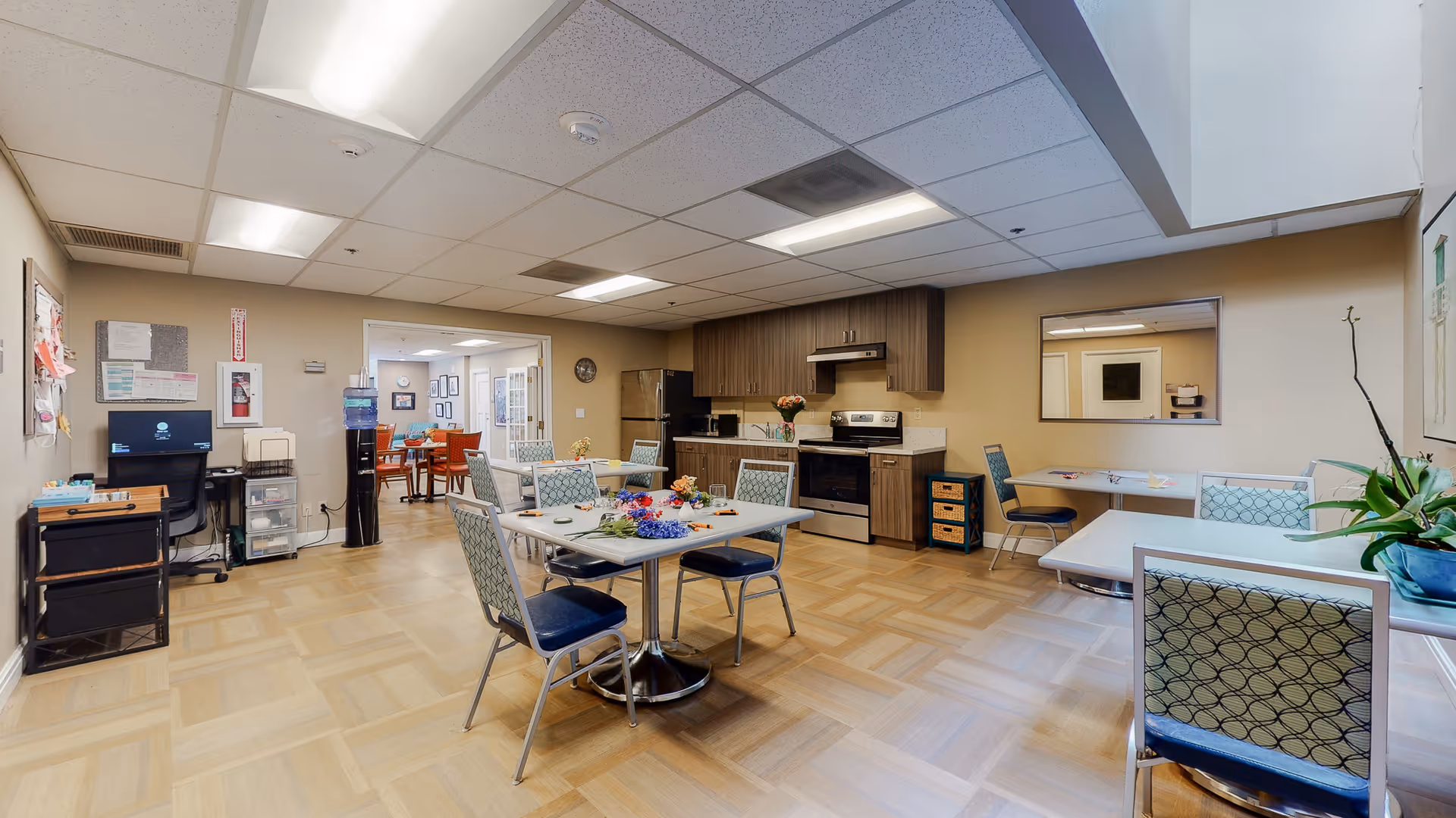 A spacious common area with a kitchenette and multiple tables with chairs arranged for seating. The kitchenette includes a stove, refrigerator, and cabinets. There is a water cooler and a small desk with a computer on the left side. The room has a light-colored floor and ceiling with fluorescent lighting. A doorway leads to another room with more seating and framed pictures on the walls.