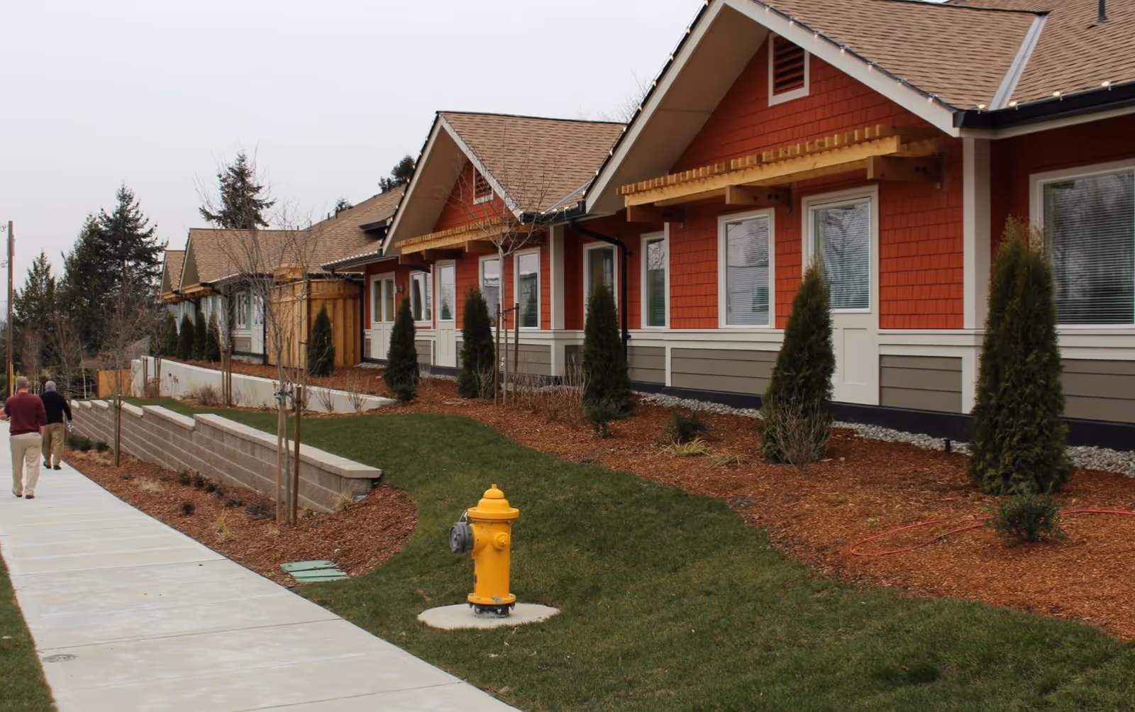 Row of single-story red cottages with landscaped beds, a sidewalk, a yellow fire hydrant, and two people walking away.