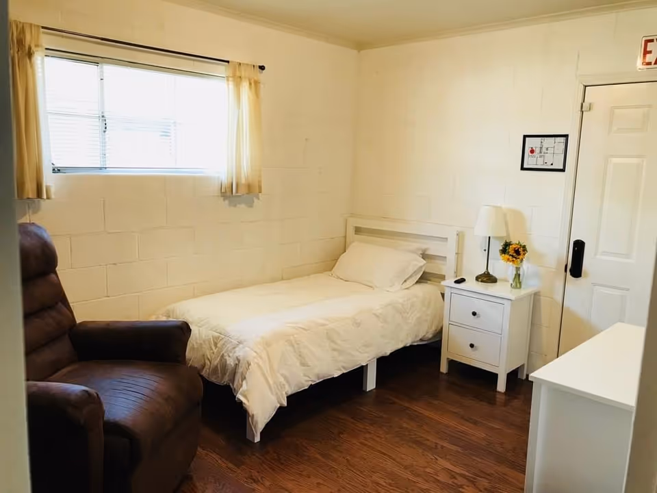 Small private bedroom with a single bed, white nightstand with a lamp and flowers, and a brown recliner by a window.
