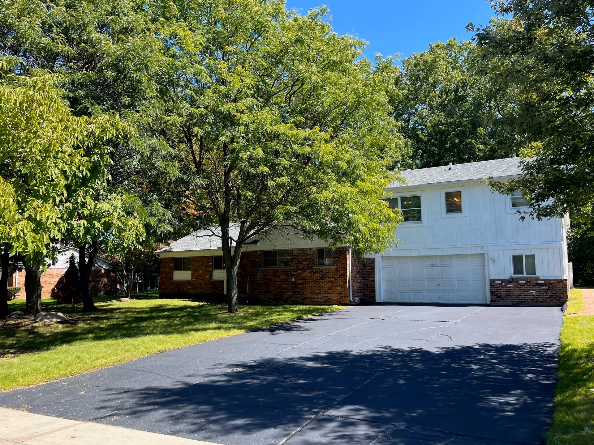 A residential-style building with a brick and white exterior surrounded by green trees and a well-maintained lawn under a clear blue sky. The building has a large driveway leading to a double garage door.