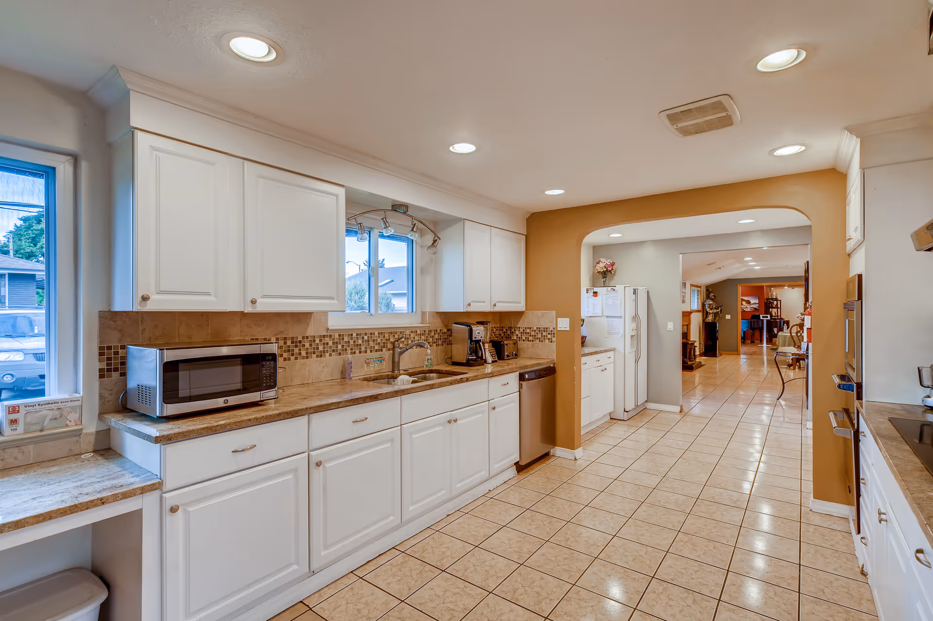A spacious kitchen with white cabinets, beige tiled floor, and recessed ceiling lights. The kitchen features a microwave, coffee maker, dishwasher, and a double sink under a window. The walls are painted white and light brown, with a tiled backsplash. An archway leads to another room with more cabinets and a refrigerator.