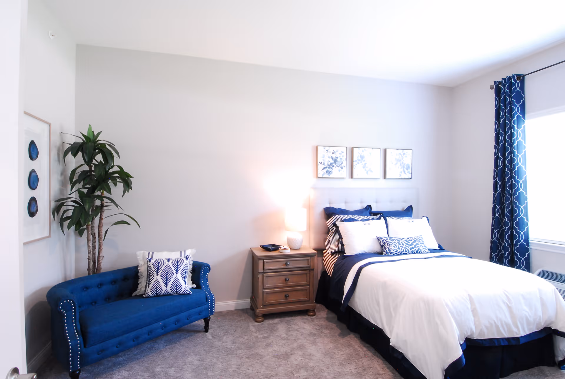 A bright bedroom with a neatly made bed featuring white and navy blue bedding, a wooden nightstand with a lamp, a blue upholstered loveseat with a decorative pillow, a tall green plant, three framed floral artworks on the wall, and a window with navy blue patterned curtains.