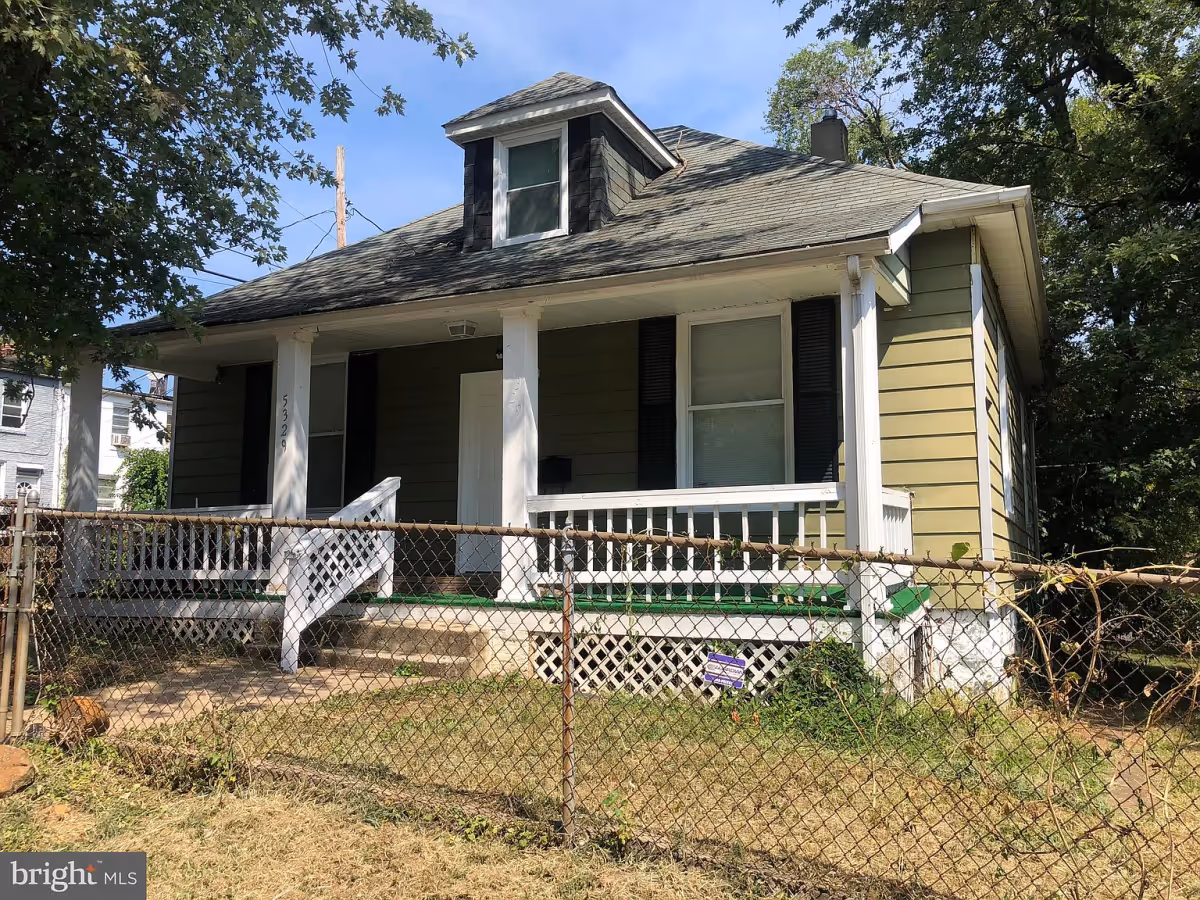 Exterior view of a small green house with a gray roof and white porch railings. The house has a front porch with steps leading up to a white door and two windows with black shutters. There is a chain-link fence in front of the house and trees surrounding the property under a clear blue sky.