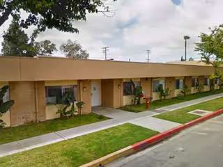 Single-story brick building with multiple doors and windows, a sidewalk, green grass, and some plants in front under a partly cloudy sky.