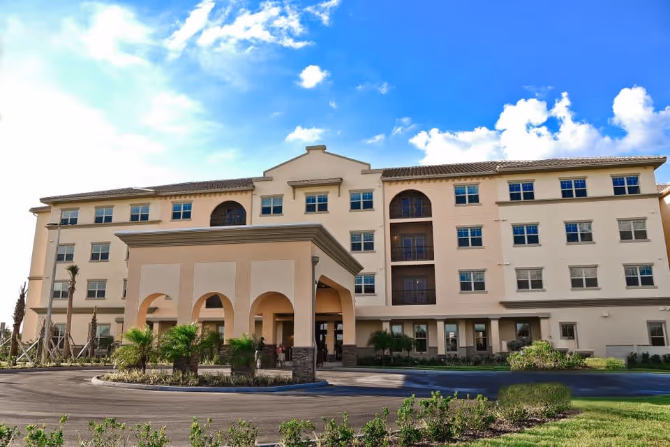 Exterior view of a multi-story senior living facility building with a covered entrance and landscaped surroundings under a bright blue sky with some clouds.