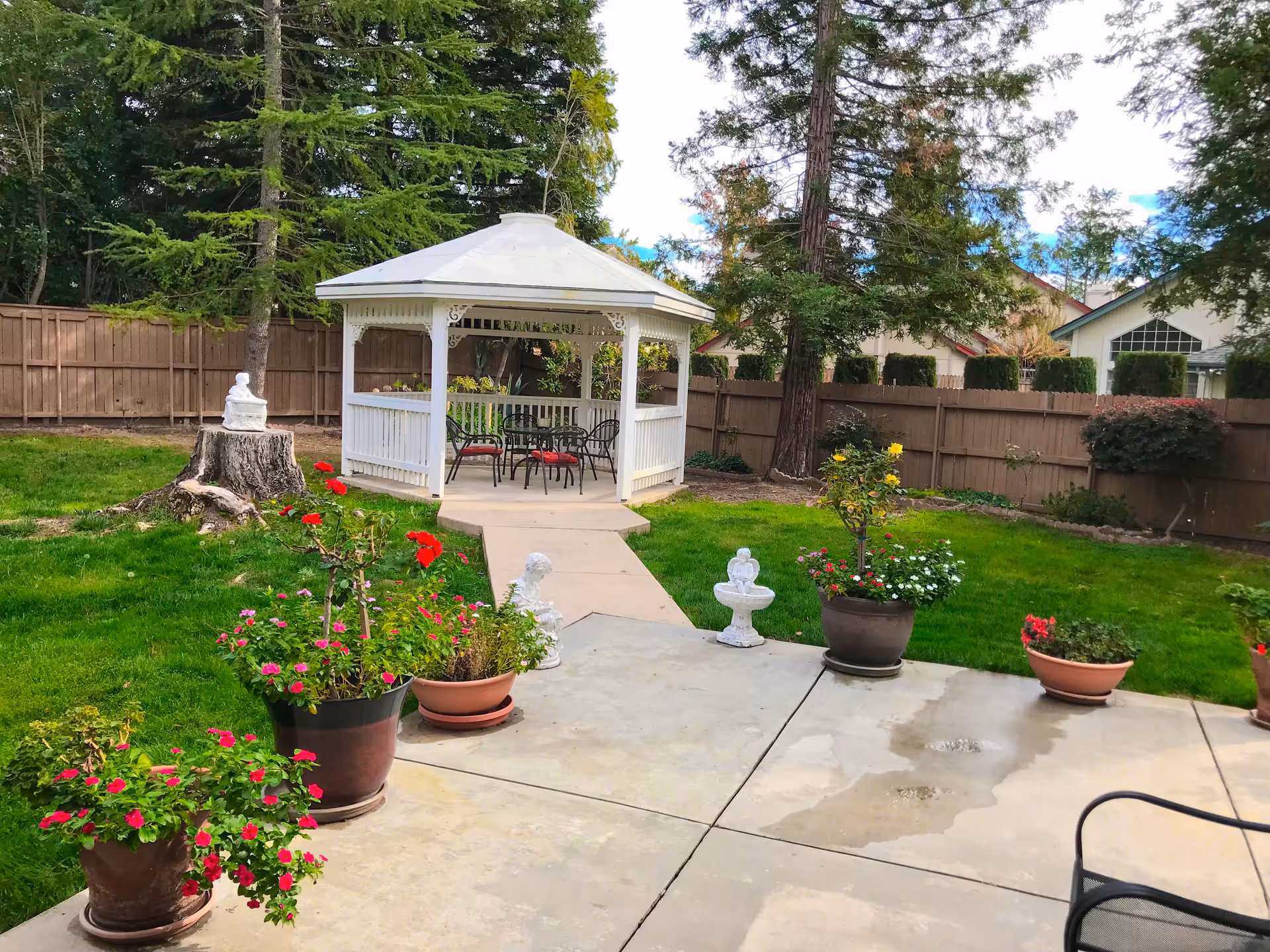 A peaceful outdoor garden area with a white gazebo containing a table and chairs. The garden has green grass, several potted flowering plants, small decorative statues, and tall trees surrounding the space. A wooden fence encloses the garden.
