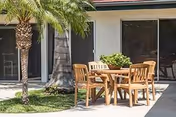 Outdoor courtyard with a wooden table and four chairs beside palm trees in front of sliding glass doors.