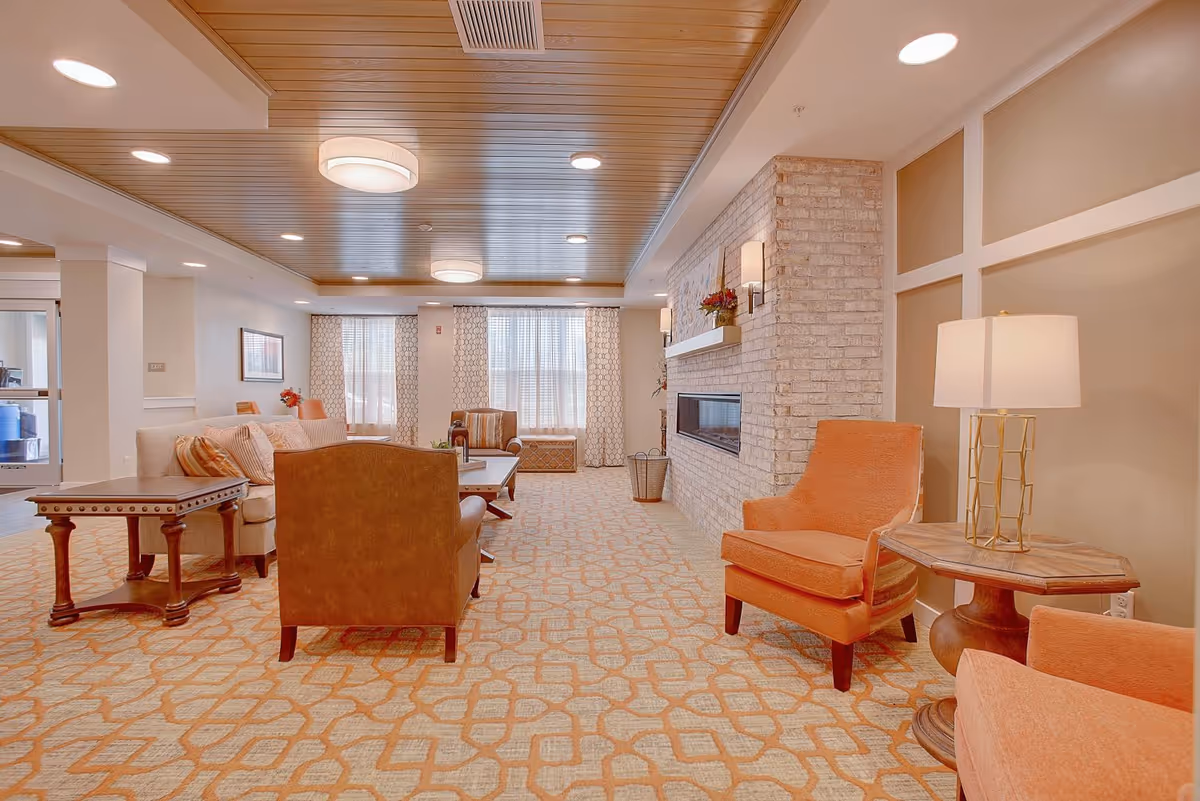 A spacious and well-lit living room area in a senior living facility with a patterned carpet, wooden ceiling, and a brick fireplace. The room is furnished with comfortable chairs in orange and brown tones, a beige sofa, wooden side tables, a coffee table, and lamps. Large windows with patterned curtains allow natural light to fill the space.