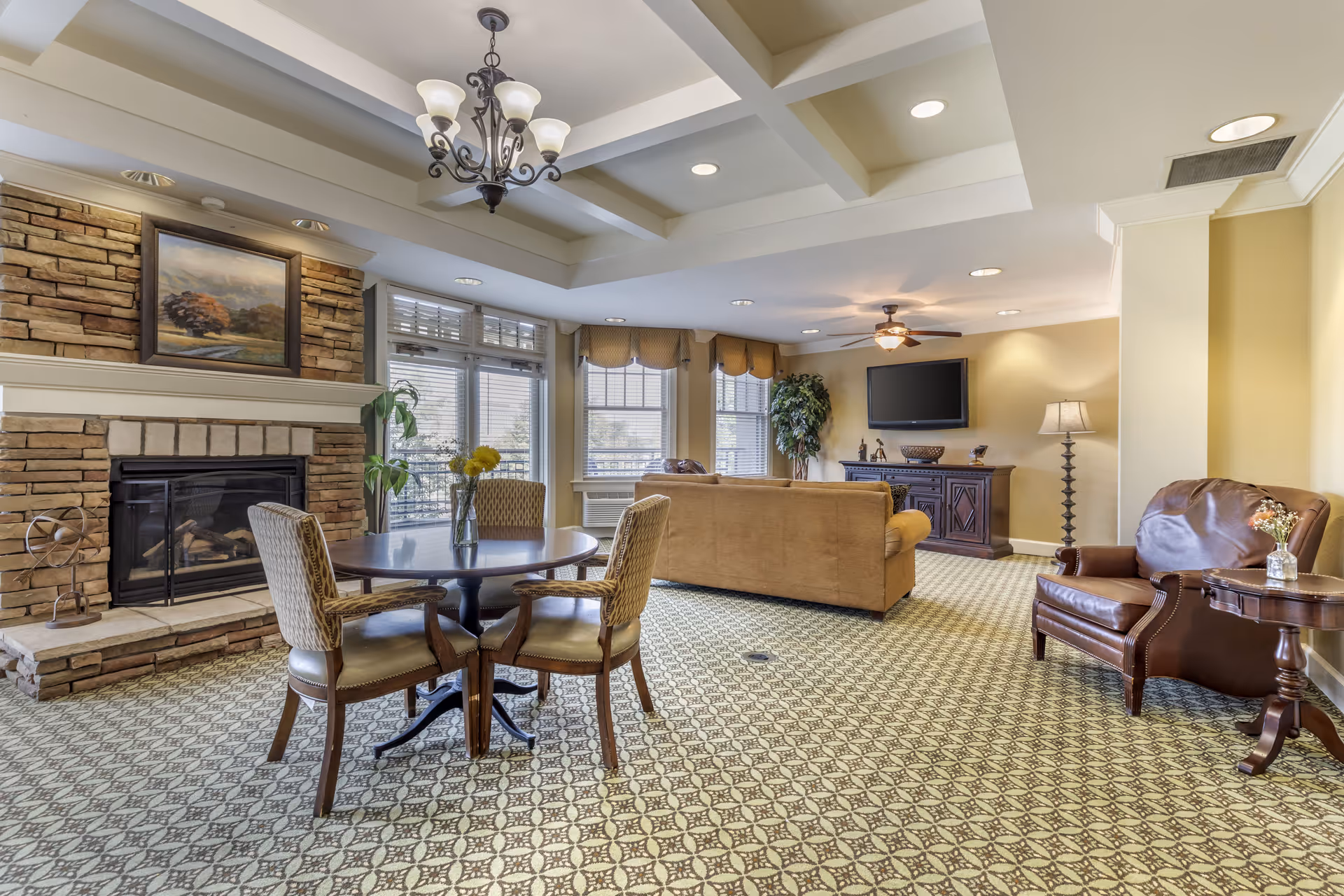 A cozy living room area in Brookdale Greenwood Village featuring a stone fireplace with a painting above it, a round wooden table with four upholstered chairs, a beige sofa facing a wall-mounted flat-screen TV, a leather armchair with a small side table and lamp, and large windows with blinds allowing natural light to fill the room.
