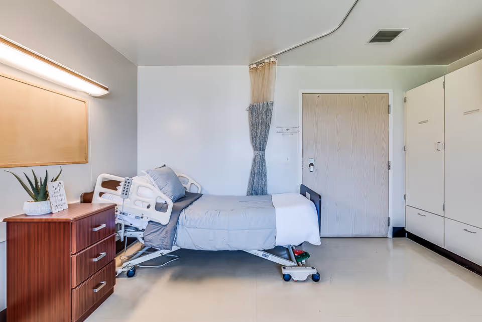 A clean and simple nursing center bedroom with a hospital bed made up with gray bedding and a white blanket. Next to the bed is a wooden nightstand with a potted plant and a small framed sign. The room has a beige door, a white wall with a curtain divider, and a large white wardrobe with multiple compartments.
