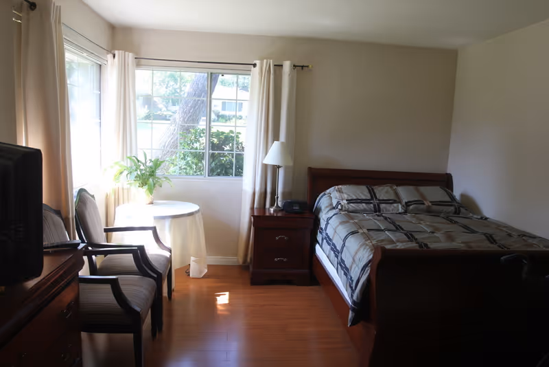 A bedroom with a wooden bed featuring a plaid comforter, a wooden nightstand with a lamp and clock, two chairs with striped cushions, a small round table with a plant, and a window with white curtains letting in natural light.
