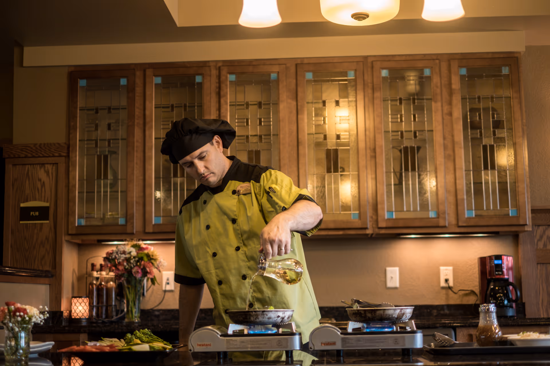 A chef wearing a green and black uniform and a black chef hat is pouring liquid from a glass pitcher into a pan on a portable stove in a kitchen with wooden cabinets and decorative glass doors. There are flowers, a coffee maker, and various kitchen items on the countertop.